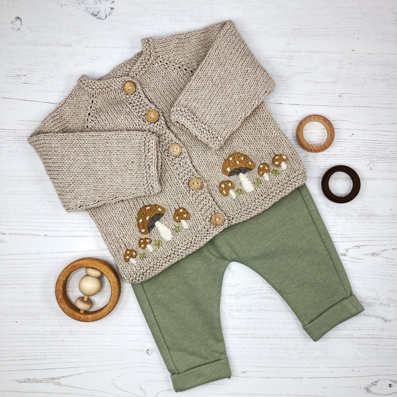 May include: A beige knitted cardigan with mushroom embroidery and a set of green pants. The cardigan has button closures and a ribbed texture.