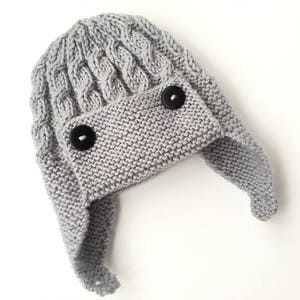 May include: Gray knitted hat with ear flaps and two black buttons. The hat has a cable knit pattern.