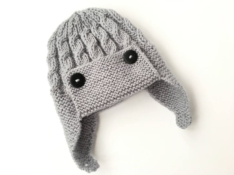 May include: Gray knitted hat with ear flaps and two black buttons. The hat has a cable knit pattern.