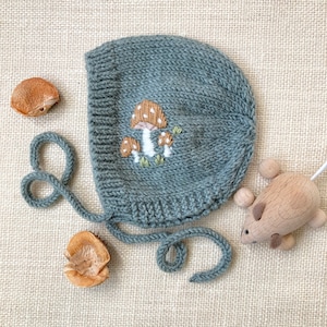 May include: A knitted baby bonnet in a muted teal color, adorned with an embroidered mushroom design. The bonnet has a rounded shape with tie closures. A wooden toy mouse and two seed pods are also in the image.