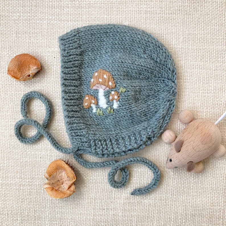 May include: A knitted baby bonnet in a muted teal color, adorned with an embroidered mushroom design. The bonnet has a rounded shape with tie closures. A wooden toy mouse and two seed pods are also in the image.