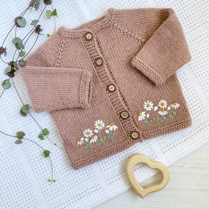 May include: A knitted baby cardigan in a light pink color with embroidered white daisies. The cardigan has a button closure and is made with a soft, textured yarn.