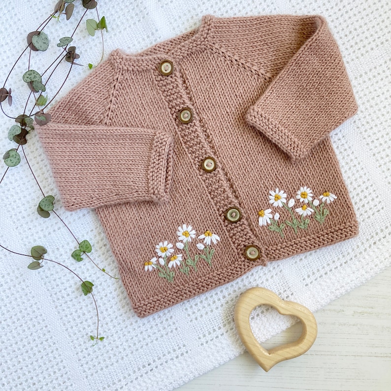 May include: A knitted baby cardigan in a light pink color with embroidered white daisies. The cardigan has a button closure and is made with a soft, textured yarn.
