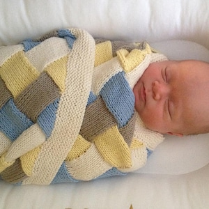 K&ouml;nnte beinhalten: Ein neugeborenes Baby ist in eine gestrickte Decke mit einem geflochtenen Muster in Blaut&ouml;nen, Gelb und Braun gewickelt. Die Decke ist aus weichem Garn gefertigt und perfekt, um ein Baby warm und gem&uuml;tlich zu halten.