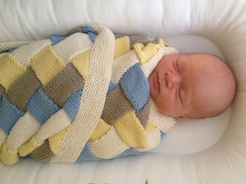 K&ouml;nnte beinhalten: Ein neugeborenes Baby ist in eine gestrickte Decke mit einem geflochtenen Muster in Blaut&ouml;nen, Gelb und Braun gewickelt. Die Decke ist aus weichem Garn gefertigt und perfekt, um ein Baby warm und gem&uuml;tlich zu halten.