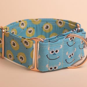 Puede incluir: Un collar de tela azul para mascotas con un patrón repetitivo de personajes circulares verdes de un solo ojo y caras de monstruos peludos azules. El collar incluye herrajes de metal en oro rosa, incluyendo una anilla en D y ajustadores.