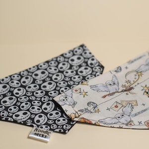 Pode incluir: Duas bandanas para cães, uma preta com rostos brancos de Jack Skellington e a outra branca com corujas e varinhas. As bandanas têm o texto "Noodle Necks" na etiqueta.
