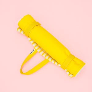 Peut inclure: Un tapis jaune vif enroulé, maintenu par deux sangles jaunes assorties et une poignée de transport jaune. L'intérieur du tapis présente un motif à carreaux blancs et jaunes. Une petite étiquette en tissu blanc avec du texte noir est fixée à la poignée.