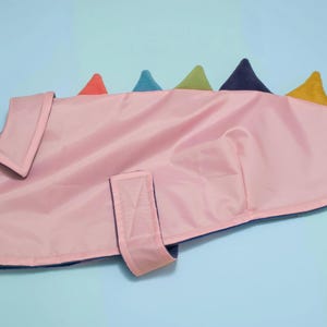May include: A light pink pet coat featuring a collar and a row of multi-coloured triangular spikes along the back. The coat includes a dark blue interior lining and a hook-and-loop fastener strap on the side for securing the garment around the torso.