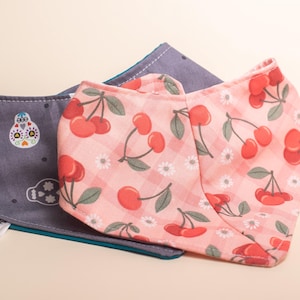 Può includere: Due bandane triangolari in tessuto con etichette bianche recanti la scritta Noodle Necks. Una bandana è grigia con un motivo a teschi, l'altra è rosa chiaro con una stampa di ciliegie rosse e margherite bianche su sfondo a quadretti.