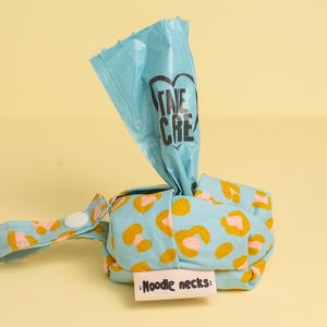 May include: A blue and yellow patterned reusable dog poop bag dispenser with a white button and a strap. The dispenser has a blue plastic bag with the text "Take Care" printed on it. The dispenser also has a tag that says "Noodle necks."