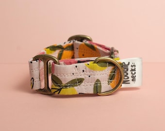 Puede incluir: Un collar de tela para mascotas con fondo rosa claro y un estampado de limones amarillos y hojas verdes. El collar incluye herrajes metálicos de color latón antiguo y una etiqueta de tela blanca con el texto negro Noodle necks: impreso.