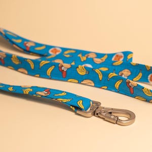 May include: A blue lanyard with a pattern of cartoon monkeys, bananas, and DK logos. The lanyard has a silver-colored metal clasp. The fabric is a bright blue color.