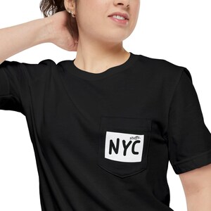 Könnte beinhalten: Schwarzes T-Shirt mit einer Tasche mit dem Text "NYC" in weißen, fetten Buchstaben auf weißem Hintergrund. Das Wort "stuffs." steht über dem Text "NYC". Das Shirt hat kurze Ärmel und einen Rundhalsausschnitt.