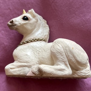 May include: A white unicorn figurine with a golden horn and a chain necklace. The unicorn is in a resting position, with its legs folded beneath it. The figurine has detailed fur texture and black eyes.