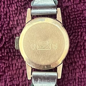 May include: A vintage gold wristwatch with a brown leather band. The watch face is round and has the markings "18K 0.750 24". The watch band is attached with silver-colored metal clasps. The watch is set against a purple background.