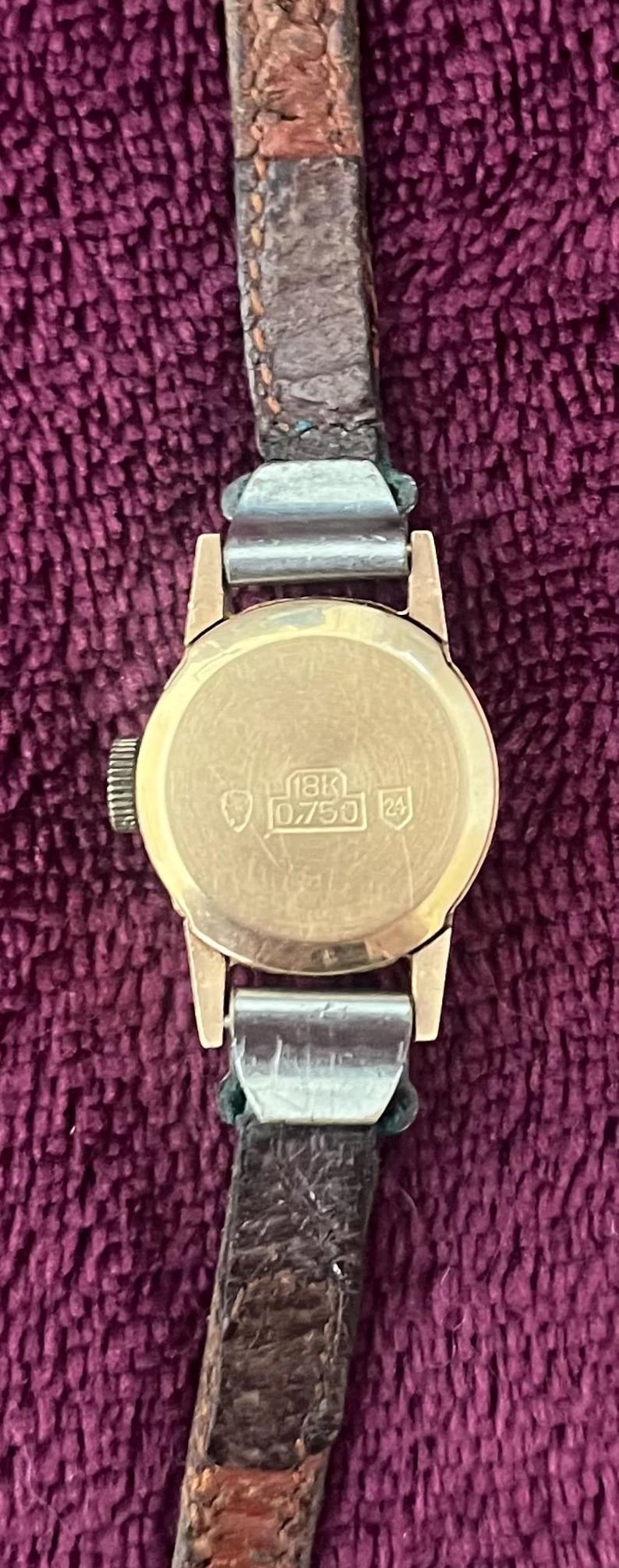 May include: A vintage gold wristwatch with a brown leather band. The watch face is round and has the markings "18K 0.750 24". The watch band is attached with silver-colored metal clasps. The watch is set against a purple background.