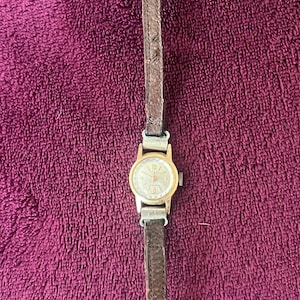 May include: A vintage wristwatch with a gold-toned face and hands, set against a brown leather band. The watch face displays numbers and markings. The band is narrow and has a buckle closure. The watch is displayed on a textured, purple surface.