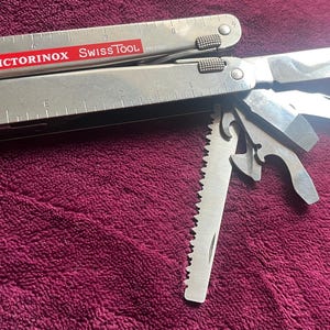 Puede incluir: Una herramienta multifunción Victorinox SwissTool plateada con una etiqueta de logotipo roja. La herramienta cuenta con una regla marcada en centímetros en el mango, una hoja principal, una hoja de sierra y otros utensilios metálicos desplegados.
