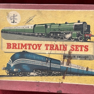 Puede incluir: Una caja rectangular de cartón para Brimtoy Train Sets con borde granate y fondo amarillo. La caja presenta ilustraciones de una locomotora de vapor verde y un tren azul aerodinámico, con el texto Brimtoy Train Sets Made in England.