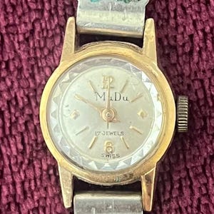 May include: A vintage gold-toned wristwatch with a round face and a brown leather band. The watch face features the text "M Du", "17 JEWELS", and "SWISS". The watch has a classic design.