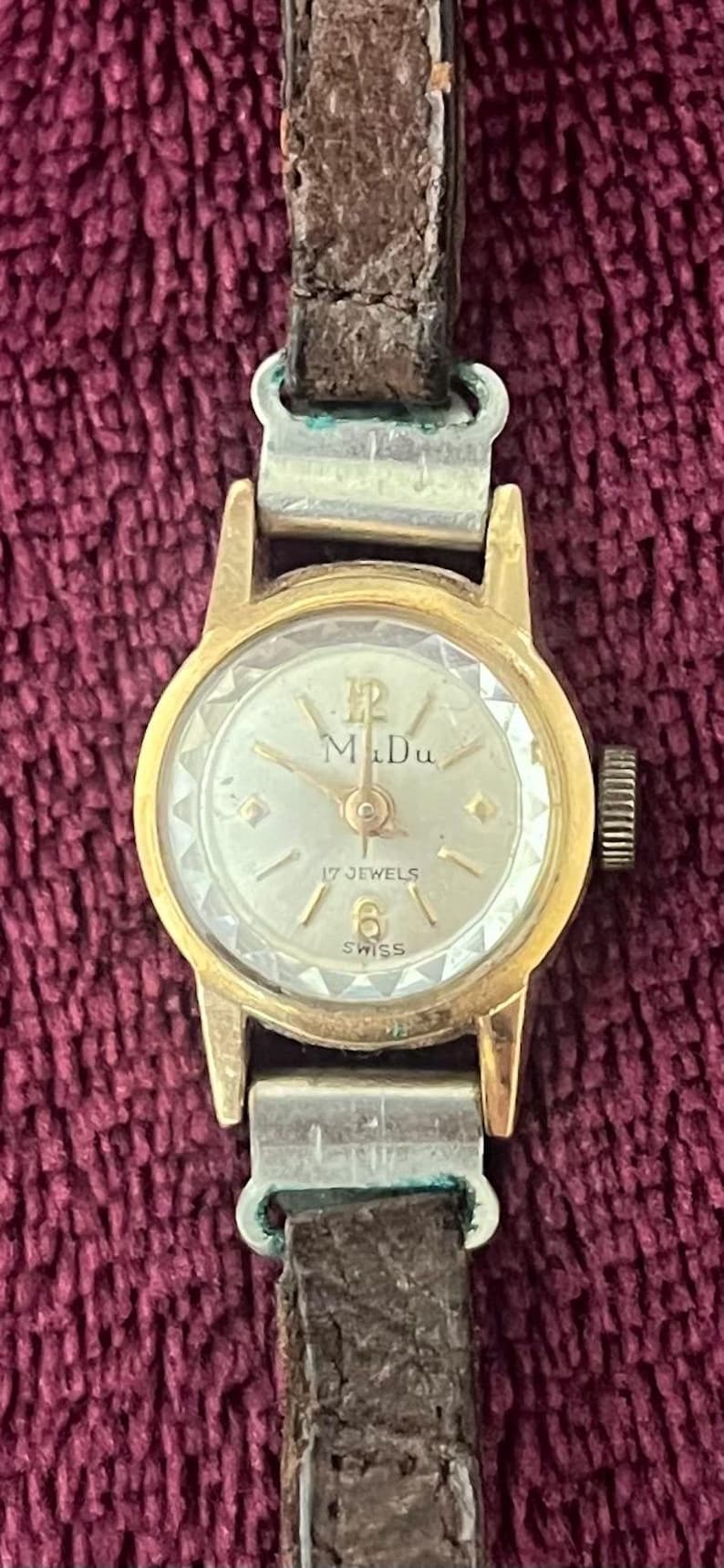 May include: A vintage gold-toned wristwatch with a round face and a brown leather band. The watch face features the text "M Du", "17 JEWELS", and "SWISS". The watch has a classic design.