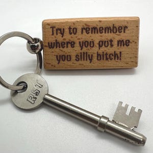 May include: A silver key and a wooden keychain. The key has the letters "RST" on the head. The rectangular wooden keychain has the engraved text "Try to remember where you put me you silly bitch!"