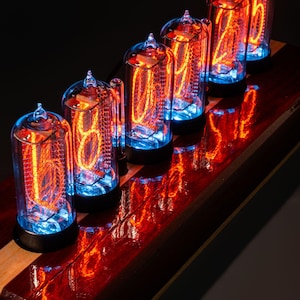 Könnte beinhalten: Sechs Vintage-Nixie-Röhren, die die Zahl 602465 anzeigen. Jede Röhre hat ein klares Glasgehäuse mit einer leuchtenden orangefarbenen Ziffer und einem blauen Sockel, montiert auf einem polierten, rotbraunen Holzsockel. Die Röhren sind vor dunklem Hintergrund beleuchtet.