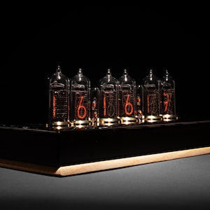 Könnte beinhalten: Ein schwarzer Holzsockel mit einer Reihe von sechs klaren Nixie-Röhren aus Glas, die die Zeit als 16:16:07 anzeigen. Die Röhren leuchten in einem warmen Orangeton.