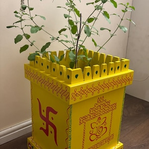 May include: A bright yellow wooden planter, adorned with red and green painted patterns. The square-shaped planter has a decorative top and contains a green plant. Religious symbols are painted on the front of the planter.