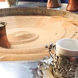Könnte beinhalten: Eine Kaffeekanne aus Kupfer mit Henkel wird verwendet, um türkischen Kaffee zuzubereiten. Die Kaffeekanne wird in eine Sandgrube gegossen. Der Sand wird verwendet, um den Kaffee abzukühlen. Auf dem Tisch stehen zwei silberne Kaffeetassen mit verzierten Designs.