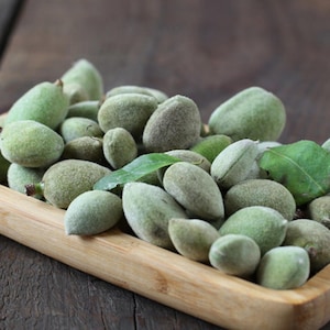 Puede incluir: Una bandeja de madera llena de almendras verdes frescas. Las almendras tienen un exterior verde claro y difuso y están agrupadas. Algunas hojas verdes se intercalan entre las almendras, y la bandeja se encuentra sobre una superficie de madera oscura.