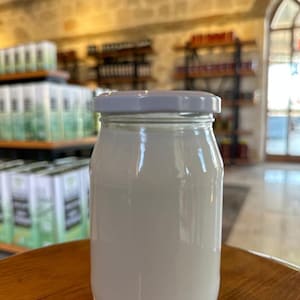 May include: A clear glass jar with a white lid, filled with a white liquid. The jar is cylindrical with a slightly rounded base. The background shows shelves with products and a stone wall.