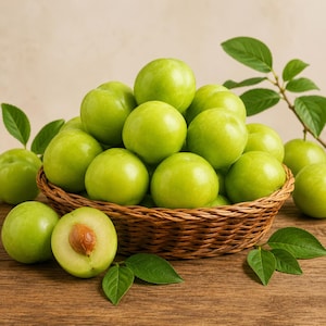 Puede incluir: Una cesta de mimbre marrón tejida llena de un montón de ciruelas frescas, redondas y de color verde claro. Una ciruela está cortada por la mitad en primer plano, revelando una pulpa clara y un solo hueso marrón. Varias hojas verdes están esparcidas en la base.