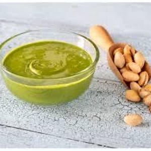 May include: A clear glass bowl filled with bright green pistachio butter sits next to a wooden scoop overflowing with shelled pistachios. The bowl and scoop are on a distressed, white-painted wooden surface.