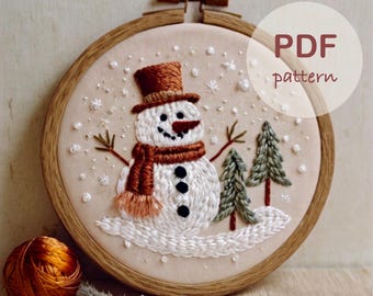 May include: Embroidered snowman with a brown hat and scarf, two green trees, and white snowflakes on a beige background in a wooden embroidery hoop. The text "PDF pattern" is in the top right corner of the image.