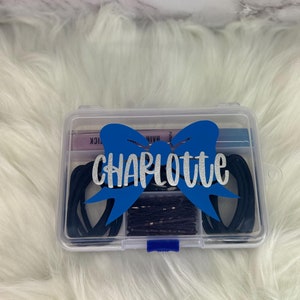 May include: A clear plastic box with a blue bow and the name "Charlotte" in silver glitter. The box contains hair accessories, including black hair ties and bobby pins. The box is on a white fluffy surface.