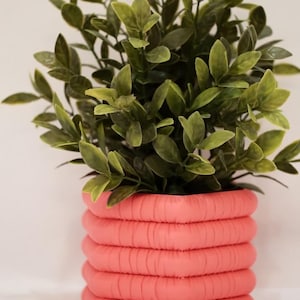 Pode incluir: Uma planta decorativa num vaso coral texturizado. O vaso tem um design cilíndrico empilhado. A planta tem várias folhas verdes. A imagem está sobre um fundo branco.