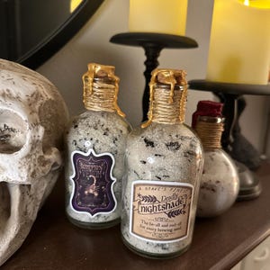 May include: Three decorative potion bottles with cork stoppers and gold wax seals. The bottles are labeled "Scorpion Venom" and "Deadly Nightshade". The bottles are filled with a granular substance and are displayed with lit candles.