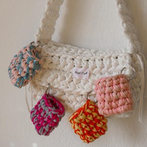 Könnte beinhalten: Eine weiße Häkeltasche mit geflochtenem Riemen und mehreren kleinen, farbenfrohen Häkelaccessoires, die mit Metallringen befestigt sind. Die Accessoires sind in Rosa-, Blau-, Orange- und Gelbtönen gehalten.