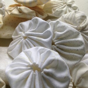 May include: Close-up of a handmade, circular textile art piece. The piece features layered, gathered fabric circles in various shades of white and cream. Some circles have a subtle shimmer, creating a textured, dimensional effect. The overall aesthetic is soft and delicate.