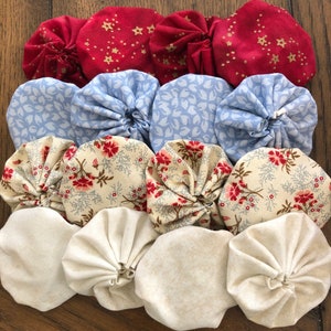 May include: An overhead shot of a collection of fabric yo-yos in various colors and patterns. The top row features red yo-yos with gold star patterns. The second row has light blue floral designs. The third row has floral patterns on a cream background. The bottom row is cream-colored.
