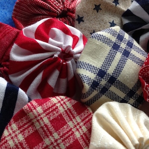 May include: A collection of fabric yo-yos in various patterns and colors. The yo-yos feature designs such as red and white stripes, blue stars on beige, and red plaid. Other colors include navy and white stripes, and a red paisley print.