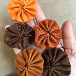 May include: Six fabric pumpkins in various shades of brown and orange. The pumpkins are all about 3 inches in diameter and have a gathered look.