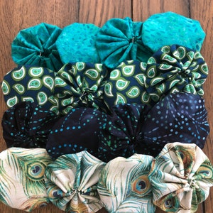 May include: A collection of handmade fabric yo-yos in various colors and patterns. The yo-yos are arranged in rows, showcasing teal, navy, and peacock feather designs. They are set against a wooden background.