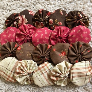 May include: A collection of fabric yo-yos in various patterns and colors. The yo-yos feature floral, plaid, and solid designs in shades of brown, red, and cream. The yo-yos are arranged in rows on a textured, light-colored surface.