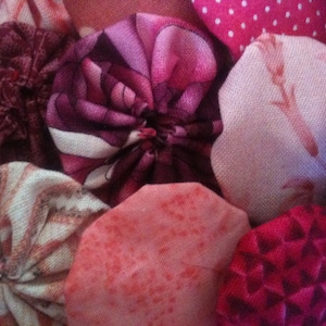 May include: Close-up of a collection of handmade fabric yo-yos in various shades of pink, red, and white. The yo-yos have different patterns, including polka dots, floral prints, and textured designs. These fabric circles are likely used for quilting or other craft projects.
