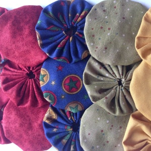 May include: Close-up of several fabric yo-yos in various colors and patterns. The yo-yos are in shades of red, blue, green, and gold. Some have floral or starburst patterns, while others are solid.