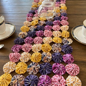 May include: A handmade table runner featuring a patchwork design of colorful fabric circles in shades of yellow, purple, and pink. A clear glass teapot sits atop the runner, with two teacups and saucers on either side.