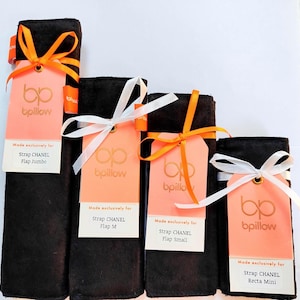 May include: Four black fabric accessories with tags. Each tag is peach-colored with the "bpillow" logo and text. The tags are tied with orange and white ribbons. The text on the tags reads "Made exclusively for Strap CHANEL Flap Jumbo, M, Small, Recta Mini."